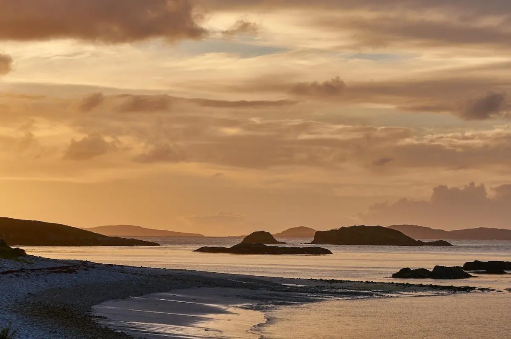 Dramatic sunset over rugged Irish west coast with rocky islands and golden sky reflecting on calm waters.